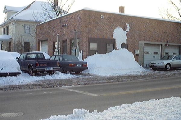 a brick building with snow piled against its wall, shaped to resemble a buffalo; several parked cars are partially covered in snow. A brick building with snow piled against its wall, shaped to resemble a buffalo; several parked cars are partially covered in snow.