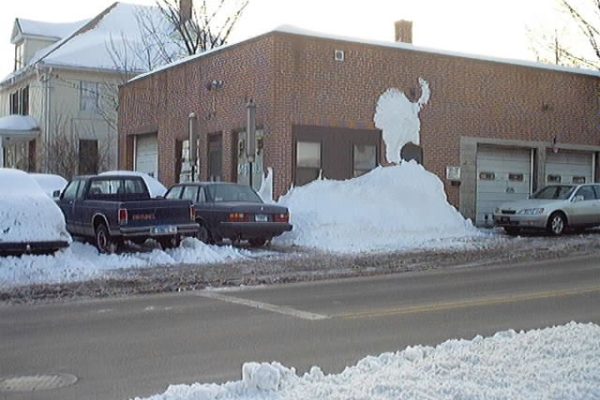 a brick building with snow piled against its wall, shaped to resemble a buffalo; several parked cars are partially covered in snow. A brick building with snow piled against its wall, shaped to resemble a buffalo; several parked cars are partially covered in snow.