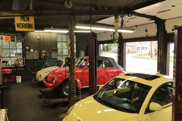 Three Volkswagen Beetle cars—two classic models in white and red, and a modern yellow one—are parked inside an auto repair garage. A “Men Working” sign hangs from the ceiling. Three Volkswagen Beetle cars—two classic models in white and red, and a modern yellow one—are parked inside an auto repair garage. A "Men Working" sign hangs from the ceiling.
