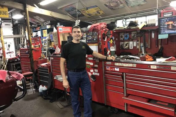 A man stands in a garage beside a large red Snap-on tool chest, surrounded by various tools and equipment under fluorescent lighting. A man stands in a garage beside a large red Snap-on tool chest, surrounded by various tools and equipment under fluorescent lighting.
