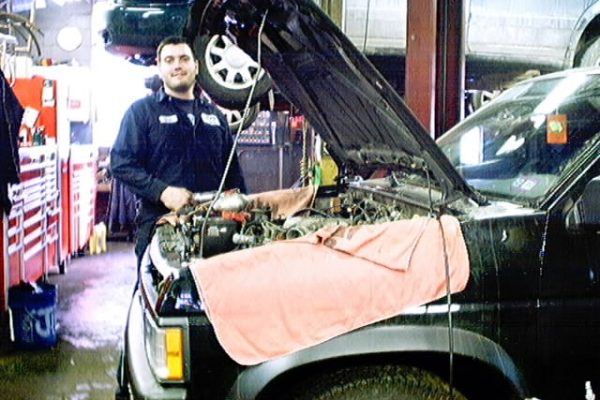 a mechanic standing next to a dark suv with the hood open and a towel draped over the fender inside an auto repair shop. A mechanic standing next to a dark SUV with the hood open and a towel draped over the fender inside an auto repair shop.
