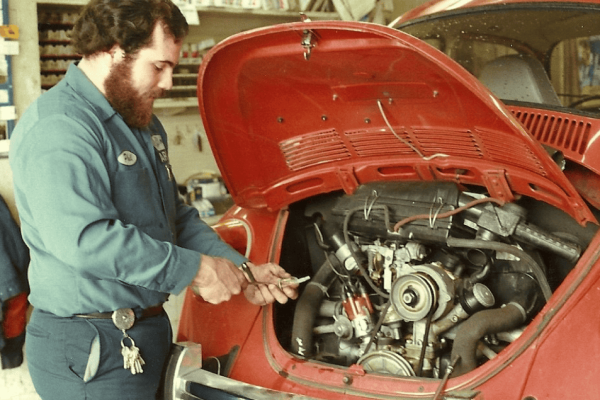 A mechanic in a blue uniform works on the engine of a red Volkswagen Beetle with the rear hood open in a garage. A mechanic in a blue uniform works on the engine of a red Volkswagen Beetle with the rear hood open in a garage.